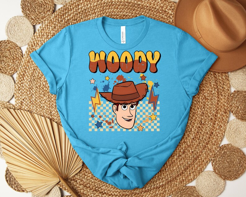 Puede incluir: Una camiseta turquesa con una ilustraci&oacute;n de dibujos animados de Woody de Toy Story. El dise&ntilde;o incluye el nombre "WOODY" en letras marrones y amarillas, una imagen de Woody, estrellas y rayos.