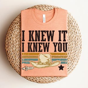 May include: A peach-colored t-shirt featuring the black text I KNEW IT I KNEW YOU above a graphic of a cowboy hat, a small rocket, and a star. The design includes horizontal stripes in muted yellow, green, and blue with a distressed texture.