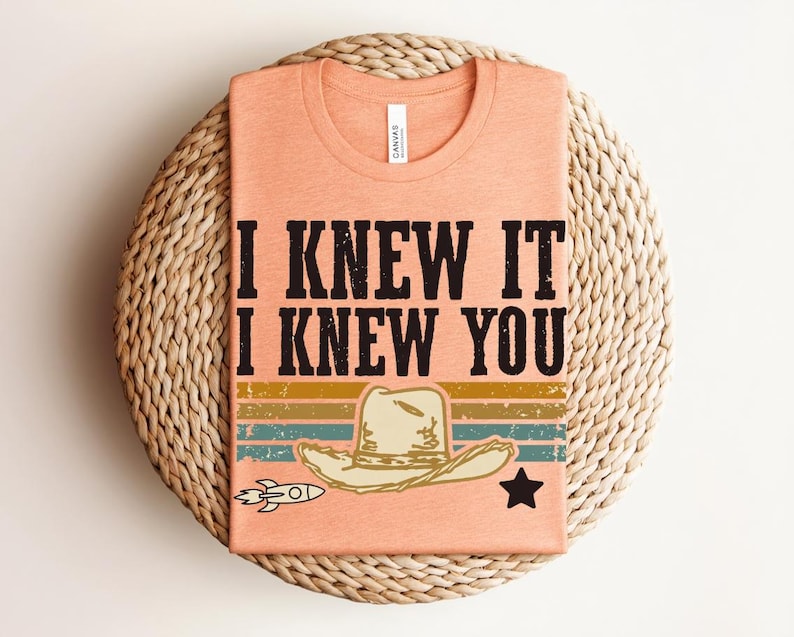 May include: A peach-colored t-shirt featuring the black text I KNEW IT I KNEW YOU above a graphic of a cowboy hat, a small rocket, and a star. The design includes horizontal stripes in muted yellow, green, and blue with a distressed texture.