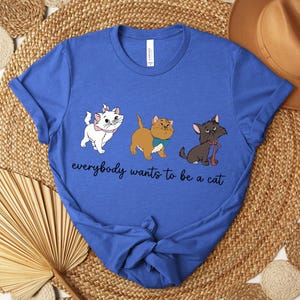 May include: Royal blue t-shirt featuring a graphic of three cartoon cats in white, brown, and black. The text "everybody wants to be a cat" is written in a cursive font below the cats. The shirt is made of a soft material.