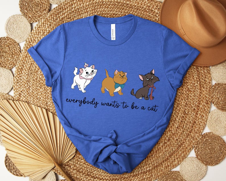 May include: Royal blue t-shirt featuring a graphic of three cartoon cats in white, brown, and black. The text "everybody wants to be a cat" is written in a cursive font below the cats. The shirt is made of a soft material.