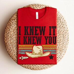 May include: A red t-shirt featuring the black text I KNEW IT I KNEW YOU above a graphic of a beige cowboy hat, a small white rocket, a black star, and horizontal stripes in muted yellow, orange, teal, and red colors across the lower section.