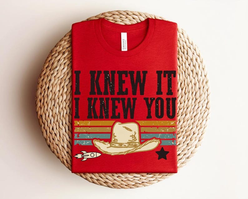 May include: A red t-shirt featuring the black text I KNEW IT I KNEW YOU above a graphic of a beige cowboy hat, a small white rocket, a black star, and horizontal stripes in muted yellow, orange, teal, and red colors across the lower section.
