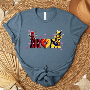 Op de afbeelding: Een blauw t-shirt met een grafisch ontwerp met het woord "MOM" in een hartvorm, met Deadpool- en Wolverine-personages. Het ontwerp bevat roze hartjes en is gedrukt op een blauw t-shirt.