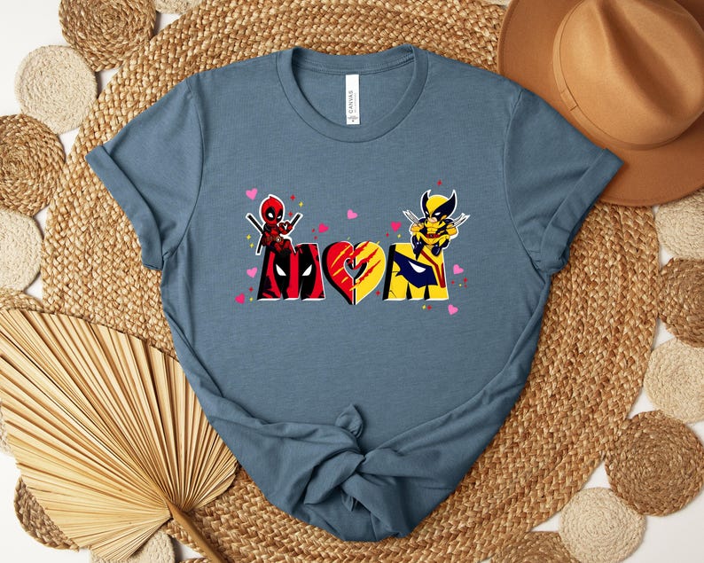 Op de afbeelding: Een blauw t-shirt met een grafisch ontwerp met het woord "MOM" in een hartvorm, met Deadpool- en Wolverine-personages. Het ontwerp bevat roze hartjes en is gedrukt op een blauw t-shirt.