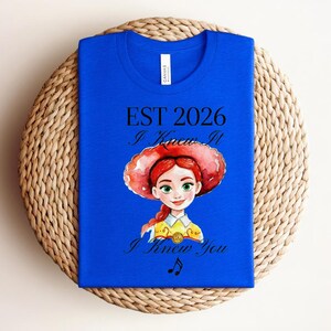 Pode incluir: Uma t-shirt azul royal com uma ilustra&ccedil;&atilde;o em estilo aguarela de uma personagem feminina com cabelo ruivo e um chap&eacute;u vermelho. O texto diz EST 2026, I Knew It e I Knew You, acompanhado por uma pequena nota musical abaixo.