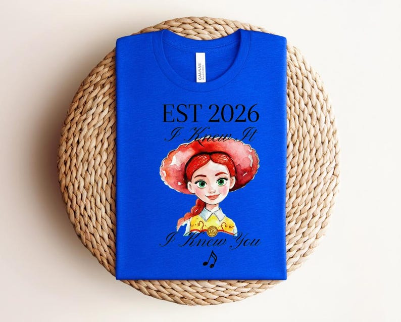 Pode incluir: Uma t-shirt azul royal com uma ilustra&ccedil;&atilde;o em estilo aguarela de uma personagem feminina com cabelo ruivo e um chap&eacute;u vermelho. O texto diz EST 2026, I Knew It e I Knew You, acompanhado por uma pequena nota musical abaixo.