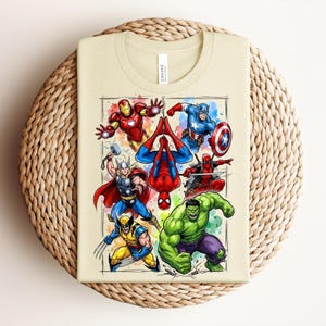May include: A beige t-shirt displaying a vibrant graphic of Marvel superheroes such as Iron Man, Captain America, and Spider-Man. The design, with a watercolor effect, features the characters in action. The t-shirt is folded on a woven, circular mat.