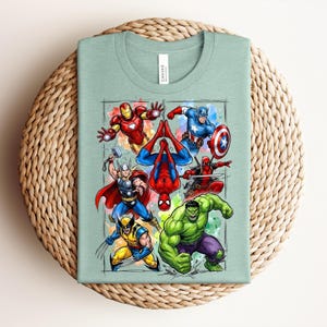May include: A pale green t-shirt with a vibrant graphic of several superheroes, such as Iron Man, Captain America, and Spider-Man. The design is set against a watercolour-style background. The shirt is folded on a woven, circular surface.