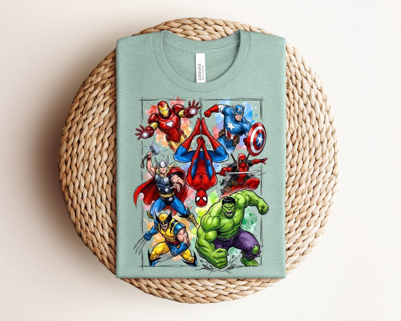 May include: A pale green t-shirt with a vibrant graphic of several superheroes, such as Iron Man, Captain America, and Spider-Man. The design is set against a watercolour-style background. The shirt is folded on a woven, circular surface.