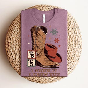May include: A mauve t-shirt featuring a graphic of a brown cowboy boot, a red cowboy hat, two stacked blocks with the text T5, and small floral and star icons. The text I KNEW IT I KNEW YOU is printed in a brown serif font at the bottom of the shirt.