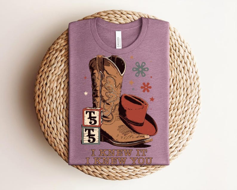 May include: A mauve t-shirt featuring a graphic of a brown cowboy boot, a red cowboy hat, two stacked blocks with the text T5, and small floral and star icons. The text I KNEW IT I KNEW YOU is printed in a brown serif font at the bottom of the shirt.