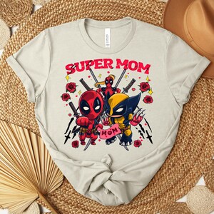K&ouml;nnte beinhalten: Hellbeige T-Shirt mit einer Grafik von Deadpool und Wolverine und dem Text "SUPER MOM". Das Design umfasst rote Rosen und Schwerter. Das T-Shirt ist aus weichem Material.