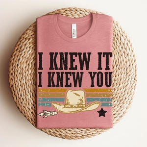 May include: A dusty rose t-shirt featuring the black text I KNEW IT I KNEW YOU. Below the text is a graphic of a beige cowboy hat, a small white rocket, and a black star, all set against horizontal stripes in yellow, teal, and orange colors.