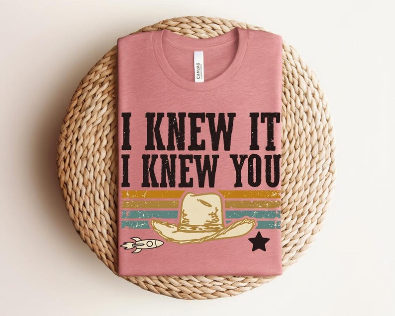 May include: A dusty rose t-shirt featuring the black text I KNEW IT I KNEW YOU. Below the text is a graphic of a beige cowboy hat, a small white rocket, and a black star, all set against horizontal stripes in yellow, teal, and orange colors.
