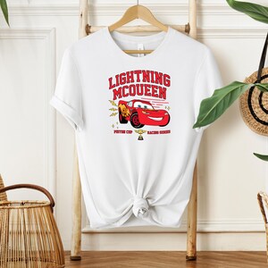 May include: White t-shirt with a red cartoon race car and the text "Lightning McQueen" in red. The car is surrounded by yellow lightning bolts and stars. The text "Piston Cup Racing Series" is below the car.