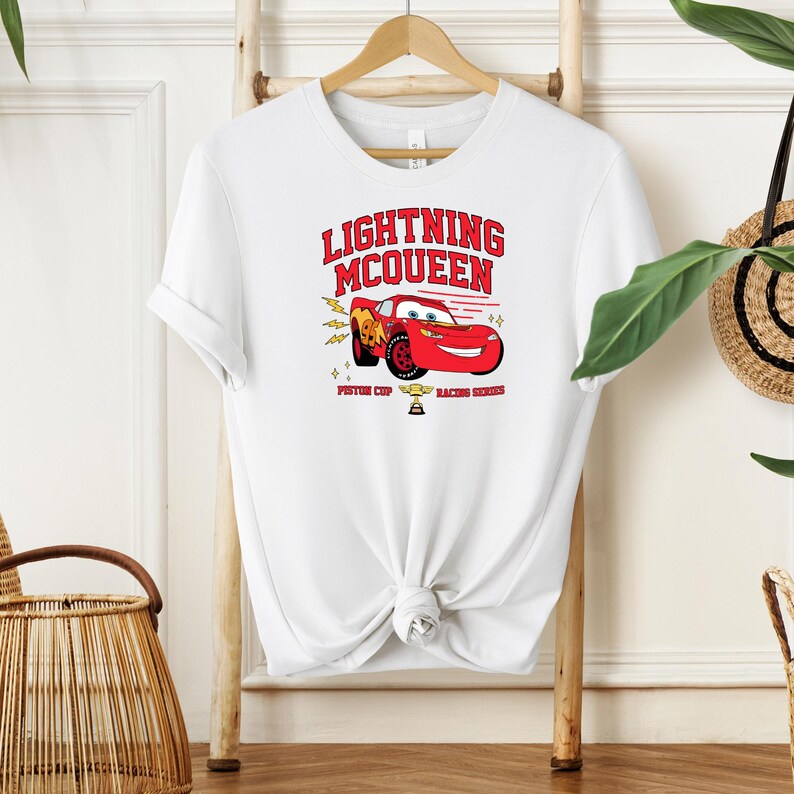 May include: White t-shirt with a red cartoon race car and the text "Lightning McQueen" in red. The car is surrounded by yellow lightning bolts and stars. The text "Piston Cup Racing Series" is below the car.
