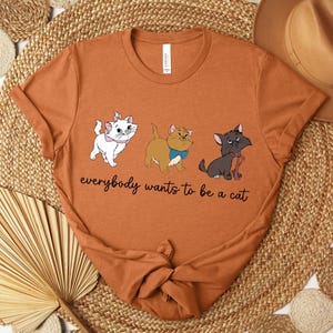 May include: Rust-colored t-shirt featuring three cartoon cats with the text "everybody wants to be a cat." The cats are white, tan, and gray, each with a unique bow or collar. The shirt is folded and placed on a woven mat.