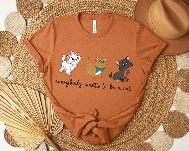 May include: Rust-colored t-shirt featuring three cartoon cats with the text "everybody wants to be a cat." The cats are white, tan, and gray, each with a unique bow or collar. The shirt is folded and placed on a woven mat.