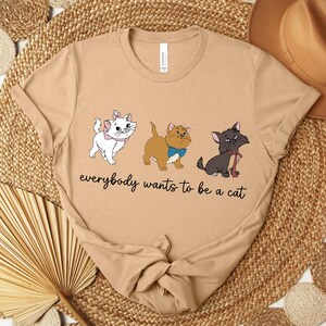 May include: Tan t-shirt featuring three cartoon cats with the text "everybody wants to be a cat". The cats are white, brown, and black, each with unique accessories. The shirt has a crew neck and short sleeves.