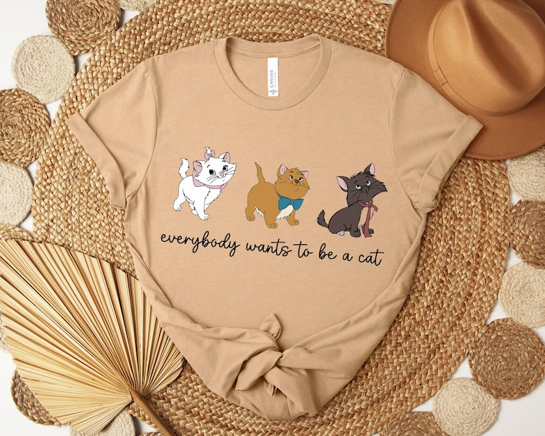 May include: Tan t-shirt featuring three cartoon cats with the text "everybody wants to be a cat". The cats are white, brown, and black, each with unique accessories. The shirt has a crew neck and short sleeves.