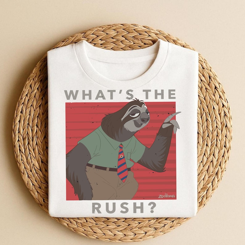 Puede incluir: Camiseta blanca con un personaje de perezoso de dibujos animados de Zootopia, con una camisa verde y una corbata a rayas rojas y azules. El texto "WHAT'S THE RUSH?" est&aacute; impreso encima y debajo de la ilustraci&oacute;n del perezoso.