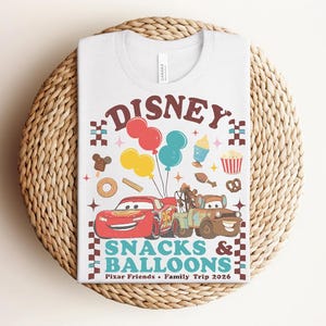 May include: White t-shirt featuring a colorful graphic with the word "Disney" above illustrations of Cars characters, balloons, and snacks. The text "Snacks & Balloons" and "Pixar Friends &bull; Family Trip 2026" are also included.
