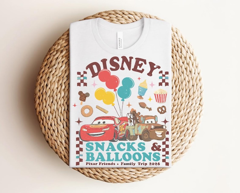 May include: White t-shirt featuring a colorful graphic with the word "Disney" above illustrations of Cars characters, balloons, and snacks. The text "Snacks & Balloons" and "Pixar Friends &bull; Family Trip 2026" are also included.