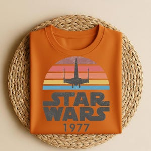 May include: Orange t-shirt featuring a retro design with the text "STAR WARS 1977" in gray. The design includes a silhouette of a spaceship against a colorful sunset. The shirt is folded on a woven, circular placemat.