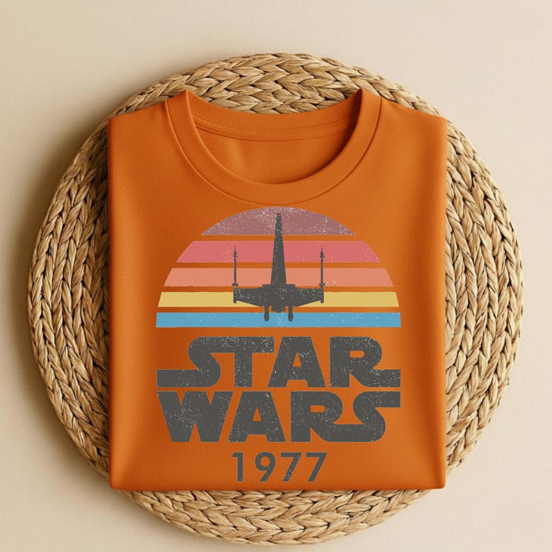 May include: Orange t-shirt featuring a retro design with the text "STAR WARS 1977" in gray. The design includes a silhouette of a spaceship against a colorful sunset. The shirt is folded on a woven, circular placemat.