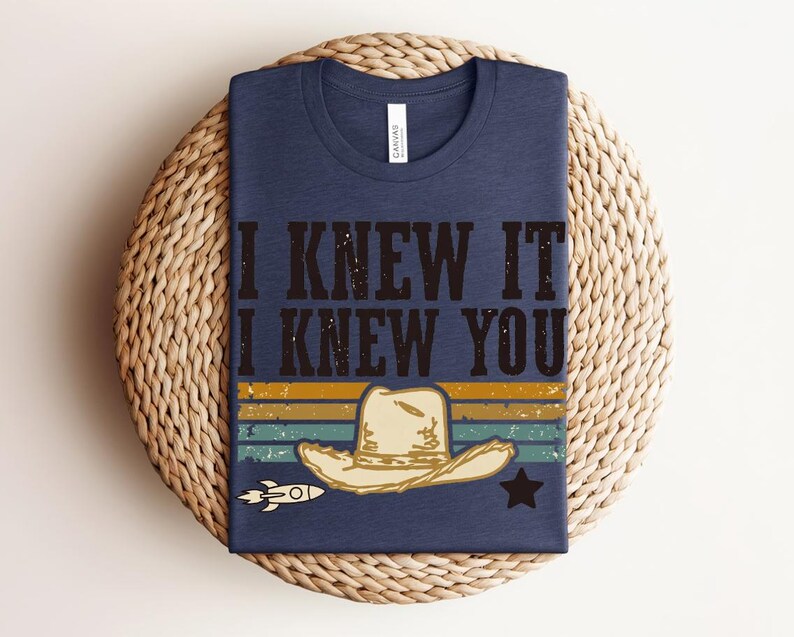 May include: A navy blue crew neck t-shirt featuring the text I KNEW IT I KNEW YOU in black distressed lettering. Below the text is a graphic of a beige cowboy hat, a small white rocket ship, and a black star over horizontal stripes.