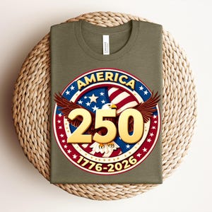 May include: An olive green t-shirt with a circular graphic featuring an eagle, the word "AMERICA", the number "250", and the years "1776-2026". The graphic is in red, white, blue, and gold.