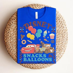 May include: A royal blue t-shirt featuring a colorful graphic design. The design includes the word "DISNEY" in brown, along with images of balloons, snacks, and the Cars characters Lightning McQueen and Mater. The text "SNACKS & BALLOONS" is also present.