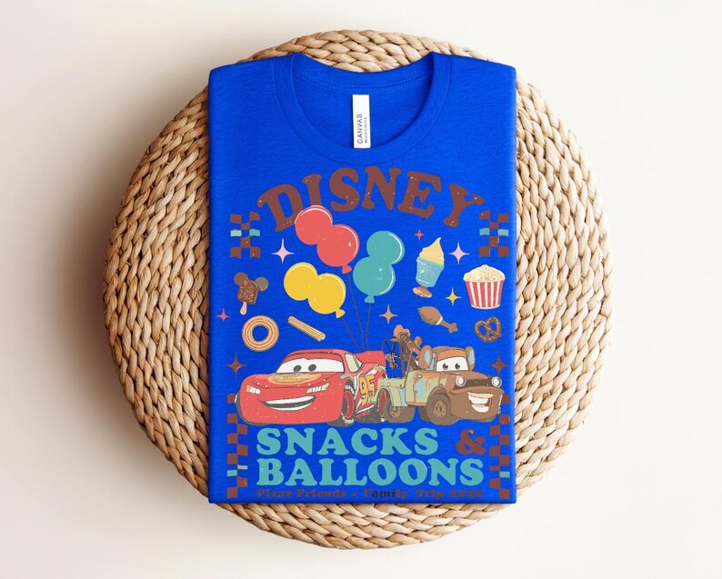 May include: A royal blue t-shirt featuring a colorful graphic design. The design includes the word "DISNEY" in brown, along with images of balloons, snacks, and the Cars characters Lightning McQueen and Mater. The text "SNACKS & BALLOONS" is also present.