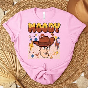 Puede incluir: Una camiseta rosa claro con una ilustraci&oacute;n de dibujos animados de Woody de Toy Story. La palabra "WOODY" est&aacute; impresa en marr&oacute;n y amarillo, con una imagen de dibujos animados de Woody con un sombrero de vaquero marr&oacute;n. Tambi&eacute;n aparecen estrellas y rayos.