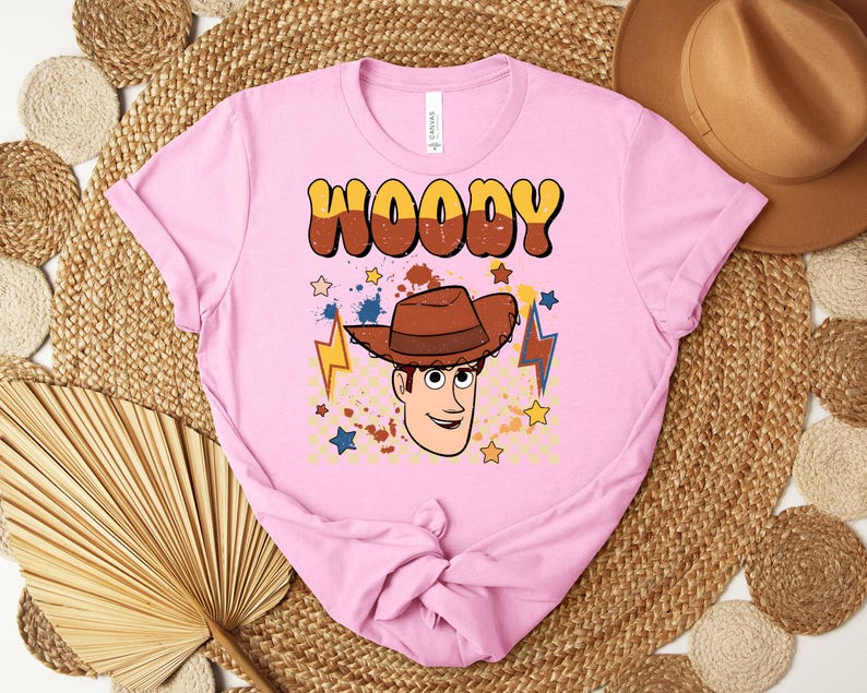 Puede incluir: Una camiseta rosa claro con una ilustraci&oacute;n de dibujos animados de Woody de Toy Story. La palabra "WOODY" est&aacute; impresa en marr&oacute;n y amarillo, con una imagen de dibujos animados de Woody con un sombrero de vaquero marr&oacute;n. Tambi&eacute;n aparecen estrellas y rayos.