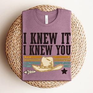 May include: A mauve short-sleeved t-shirt featuring the black text I KNEW IT I KNEW YOU above a graphic of a beige cowboy hat, a small rocket ship, and a black star, set against horizontal stripes in muted yellow, teal, and orange colors.