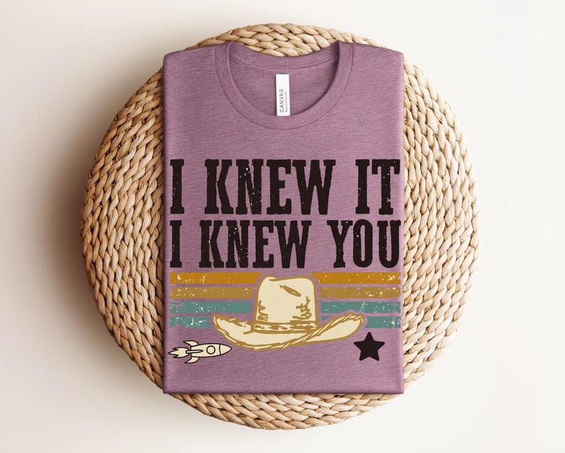 May include: A mauve short-sleeved t-shirt featuring the black text I KNEW IT I KNEW YOU above a graphic of a beige cowboy hat, a small rocket ship, and a black star, set against horizontal stripes in muted yellow, teal, and orange colors.