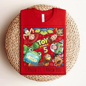 K&ouml;nnte beinhalten: Ein rotes T-Shirt mit einem Grafikdruck von Toy Story-Figuren, darunter Woody, Buzz Lightyear, Rex, Slinky Dog, Mr. Potato Head und gr&uuml;ne Aliens. Der Text lautet Toy Story 5 und I Knew It, I Knew You. Das Shirt hat einen Rundhalsausschnitt.