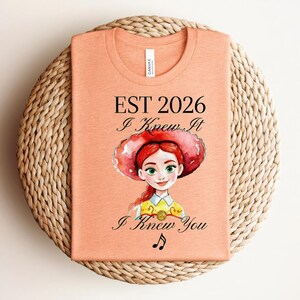 Pode incluir: Uma t-shirt cor de p&ecirc;ssego com uma ilustra&ccedil;&atilde;o em estilo aguarela de uma personagem ruiva com um chap&eacute;u vermelho. O texto diz EST 2026, I Knew It e I Knew You, acompanhado por um pequeno s&iacute;mbolo de nota musical centrado abaixo do gr&aacute;fico.