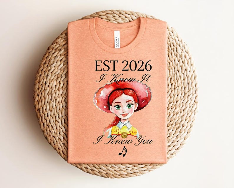 Pode incluir: Uma t-shirt cor de p&ecirc;ssego com uma ilustra&ccedil;&atilde;o em estilo aguarela de uma personagem ruiva com um chap&eacute;u vermelho. O texto diz EST 2026, I Knew It e I Knew You, acompanhado por um pequeno s&iacute;mbolo de nota musical centrado abaixo do gr&aacute;fico.
