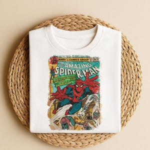 May include: White t-shirt featuring a vintage comic book cover design. The graphic showcases Spider-Man in action, with the title "The Amazing Spider-Man" prominently displayed. The design includes text that reads "Marvel Comics Group".