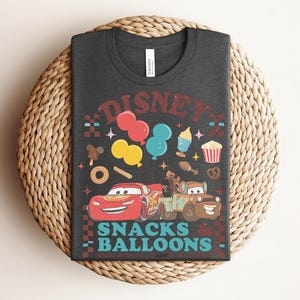 May include: Dark gray t-shirt featuring a colorful graphic design with the text "DISNEY SNACKS & BALLOONS". The design includes cartoon cars, balloons, and various snacks. The shirt is laid on a woven surface.