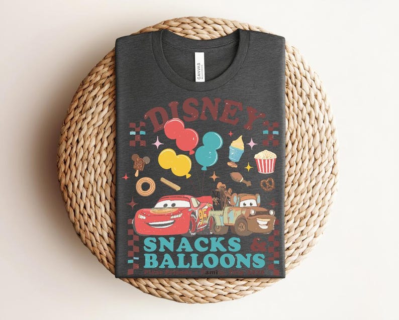 May include: Dark gray t-shirt featuring a colorful graphic design with the text "DISNEY SNACKS & BALLOONS". The design includes cartoon cars, balloons, and various snacks. The shirt is laid on a woven surface.