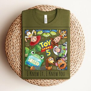 K&ouml;nnte beinhalten: Ein olivgr&uuml;nes T-Shirt mit einem Grafikdruck von Toy Story-Figuren, darunter Woody, Buzz Lightyear, Jessie, Rex, Slinky Dog, Mr. Potato Head und Aliens. Der Text lautet Toy Story 5 und I Knew It, I Knew You in schwarzer Schrift.