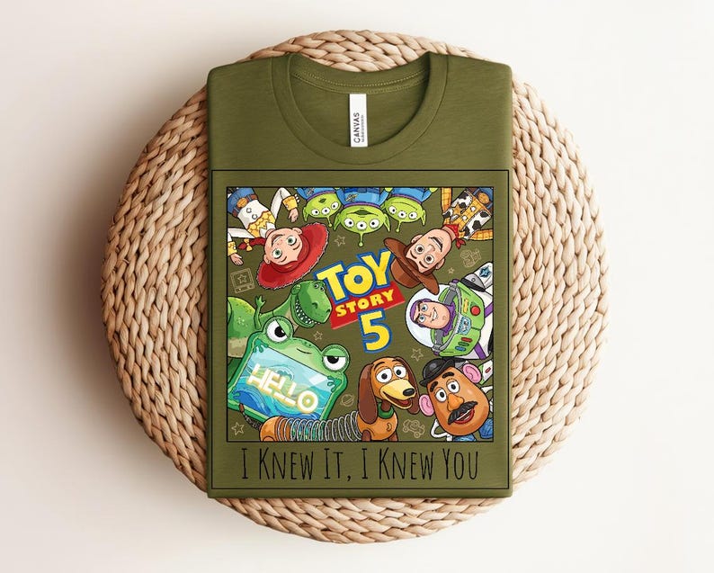 K&ouml;nnte beinhalten: Ein olivgr&uuml;nes T-Shirt mit einem Grafikdruck von Toy Story-Figuren, darunter Woody, Buzz Lightyear, Jessie, Rex, Slinky Dog, Mr. Potato Head und Aliens. Der Text lautet Toy Story 5 und I Knew It, I Knew You in schwarzer Schrift.