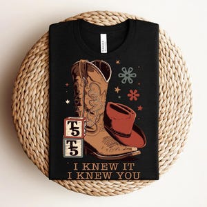 May include: A black t-shirt featuring a graphic of a brown cowboy boot, a red cowboy hat, two stacked blocks with the text T5, and small star and flower icons. The text I KNEW IT I KNEW YOU is printed in a tan font at the bottom of the graphic.