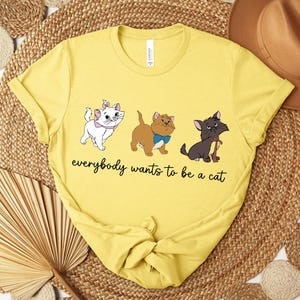 May include: Yellow t-shirt featuring three cartoon cats in different colors. The white cat has a pink bow, the brown cat has a blue bow, and the black cat has a red scarf. The text "everybody wants to be a cat" is written below the cats.