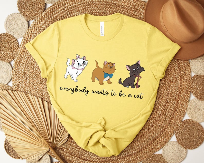 May include: Yellow t-shirt featuring three cartoon cats in different colors. The white cat has a pink bow, the brown cat has a blue bow, and the black cat has a red scarf. The text "everybody wants to be a cat" is written below the cats.