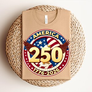 May include: A folded tan t-shirt with a circular graphic. The graphic features the word "AMERICA" in blue, a red and white striped background, stars, and the number "250" in gold. The design includes an eagle and the dates "1776-2026".
